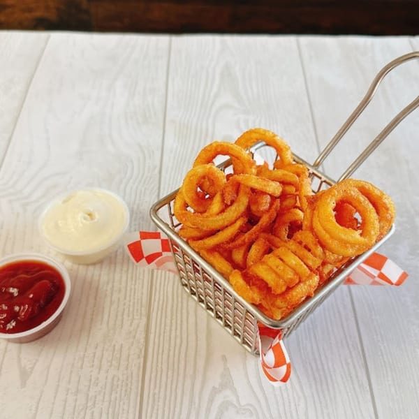 A metal basket of crispy curly fries with small cups of ketchup and mayonnaise on a white wooden table.